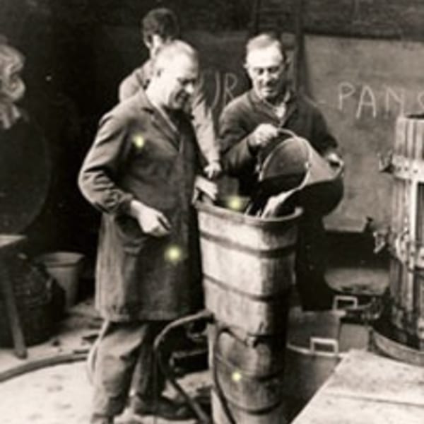 Two men in aprons pour liquid into a large barrel, possibly making wine or olive oil.