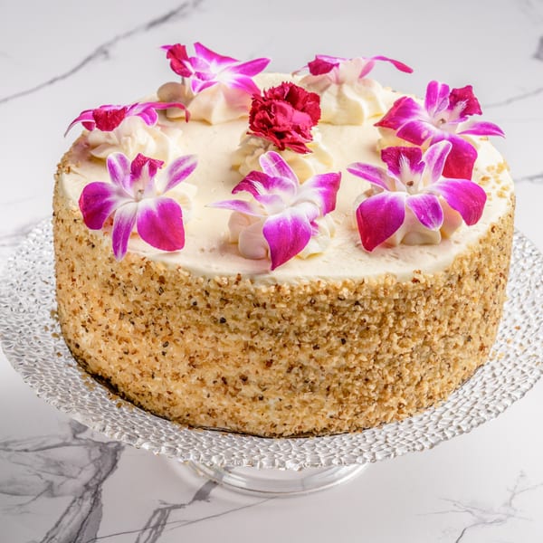 A round cake with white frosting, decorated with pink orchids and a red carnation, on a glass stand.