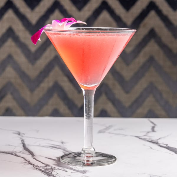 A vibrant pink cocktail in a martini glass, garnished with a delicate purple orchid, on a marble surface.
