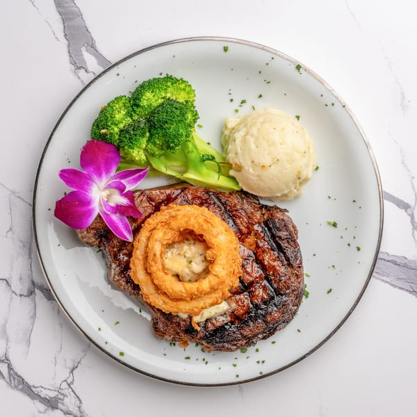 Grilled steak topped with onion rings, served with mashed potatoes, broccoli, and a purple orchid.