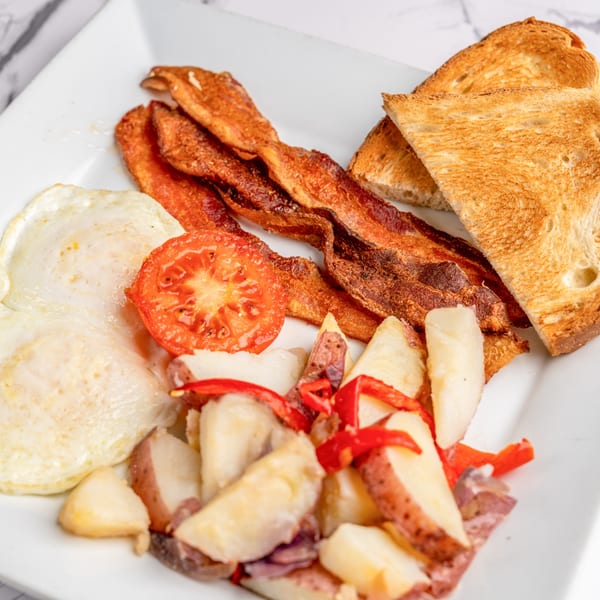Breakfast plate with crispy bacon, fried eggs, roasted potatoes, grilled tomato, and toast.